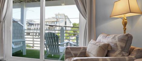 A room with a sofa, a lamp, and a view through a sliding glass door to a balcony with a chair and a house in the background.