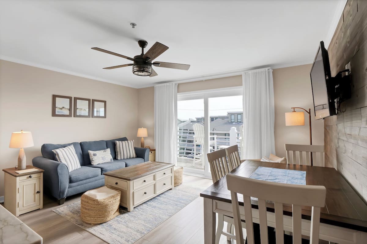 Un salon moderne avec un canapé, une table basse et une table à manger. On y trouve un ventilateur de plafond, un téléviseur fixé au mur et une fenêtre avec des rideaux.