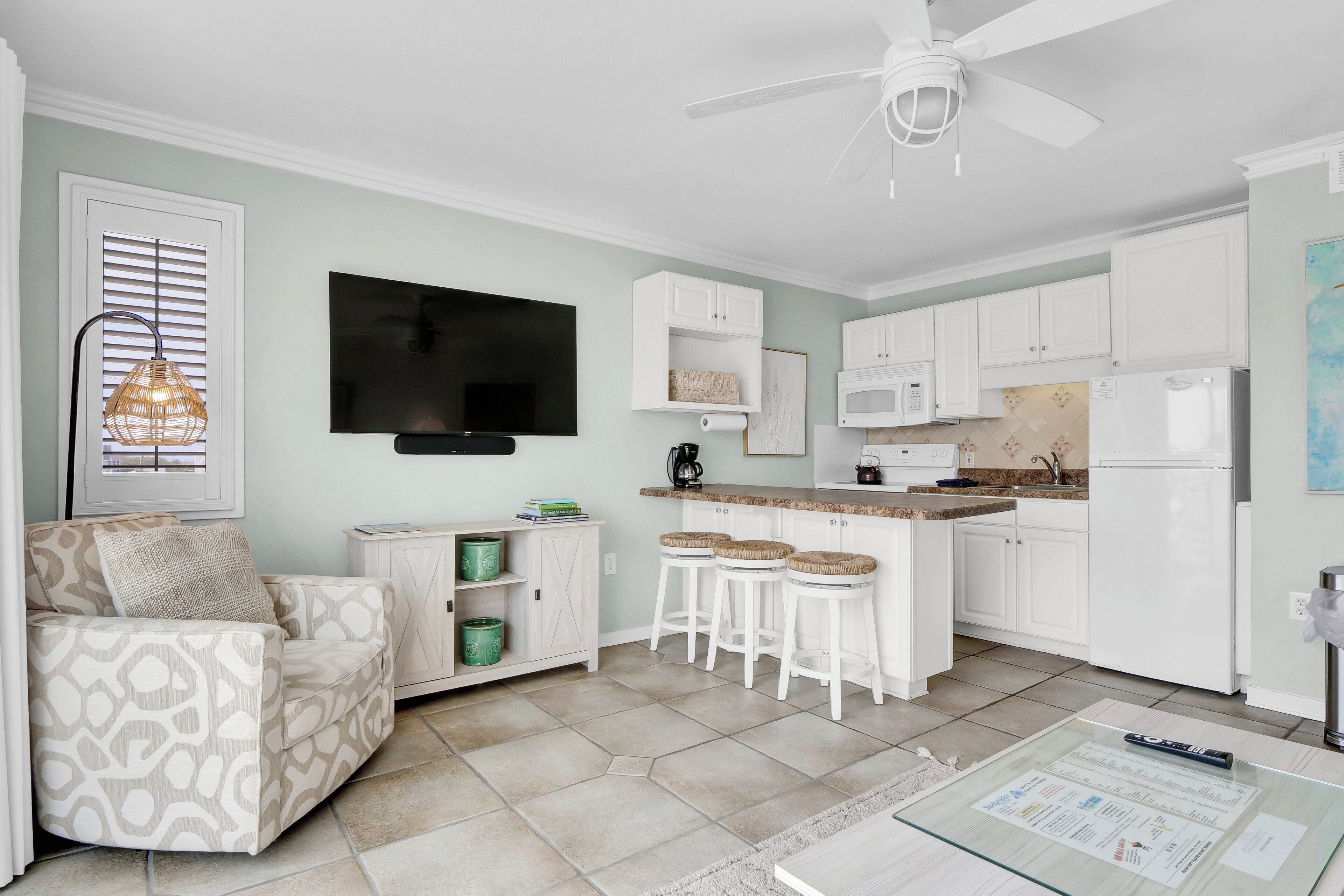 A compact kitchen with white cabinets, a microwave, and a refrigerator. There is a bar stool set and a ceiling fan.