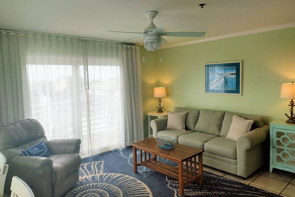 Un salon avec un ventilateur de plafond, un canapé, un fauteuil, une table basse et une lampe.