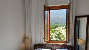 Cama bem arrumada com lençóis e toalhas brancas, um ventilador de teto, janela com vista para o verde e mesa de cabeceira com abajur.
