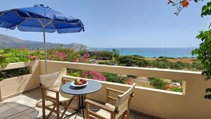 Uma varanda com mesa e cadeiras, um guarda-sol azul, e vista para o mar e montanhas.