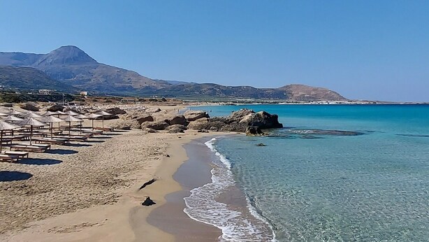 Uma praia com águas cristalinas, um primeiro plano rochoso e uma cordilheira ao longe.