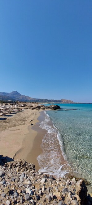 A beach with clear water, a rocky foreground, and a distant mountain range. - Doma Apartments (Kissamos)
