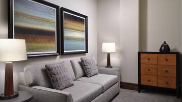 A modern living room with a grey sofa, two framed abstract paintings, a side table with a lamp, a wooden cabinet with drawers, and a patterned rug.