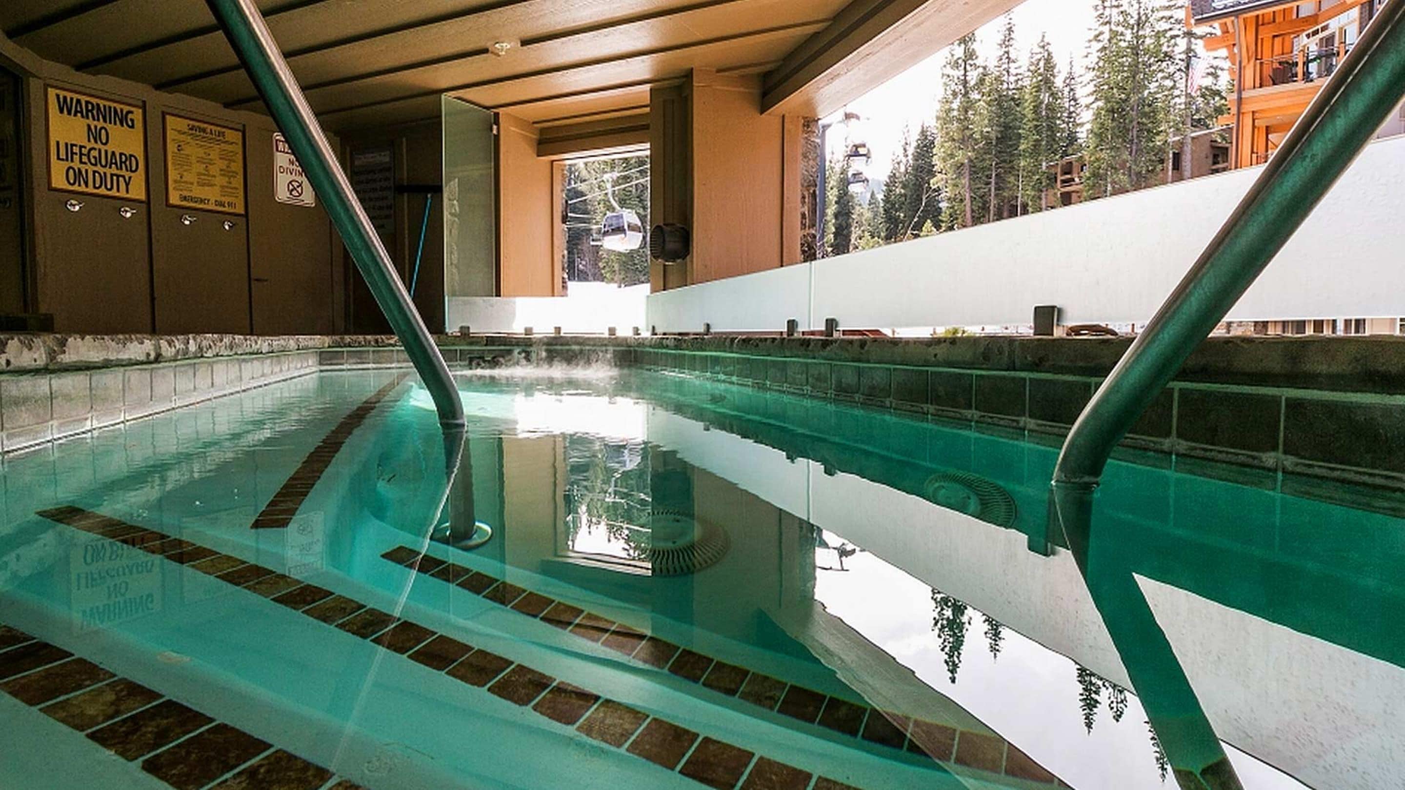 Una piscina interior con agua cristalina, un letrero de advertencia de salvavidas y vistas a árboles y un edificio en el exterior.