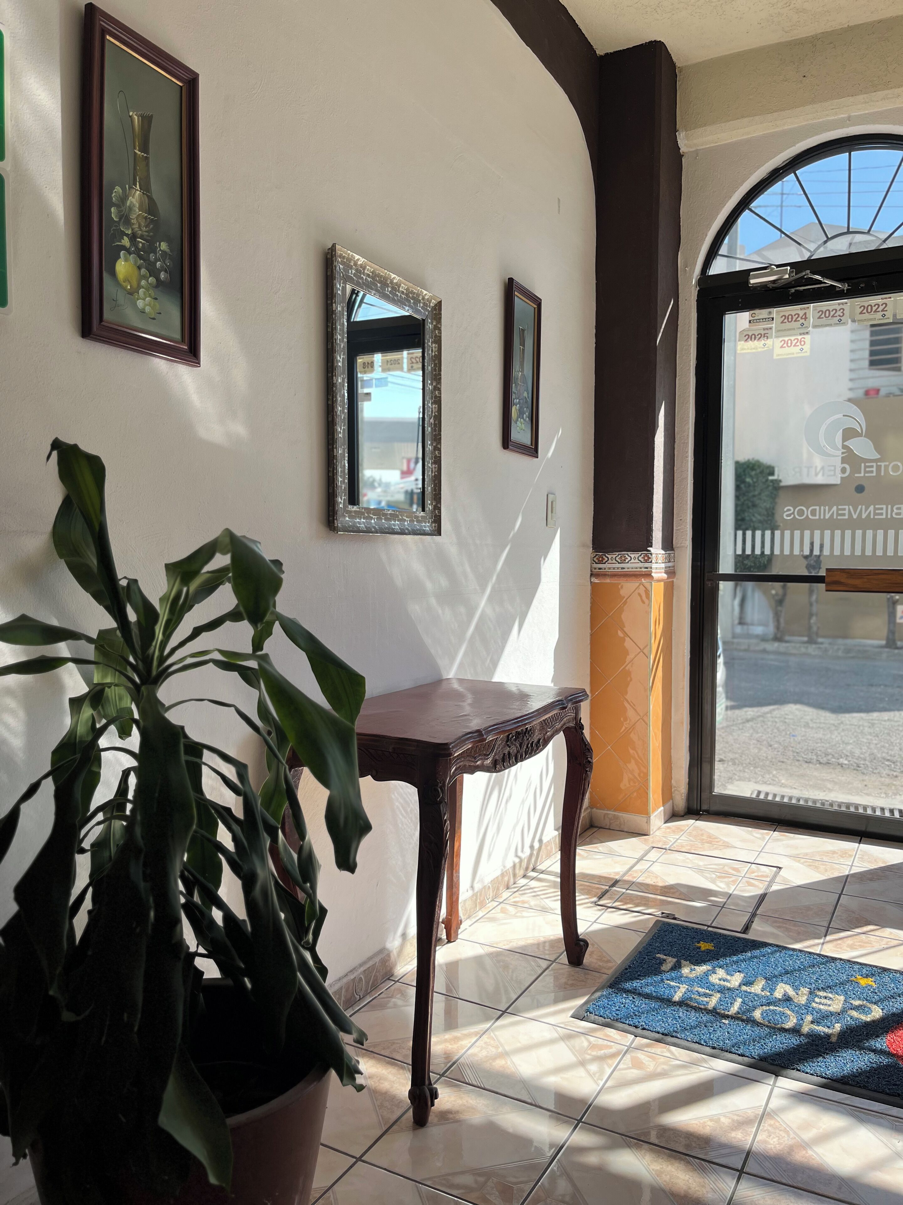 A hotel lobby with a potted plant, a wooden table, framed artwork on the wall, and a doormat with the hotel name.