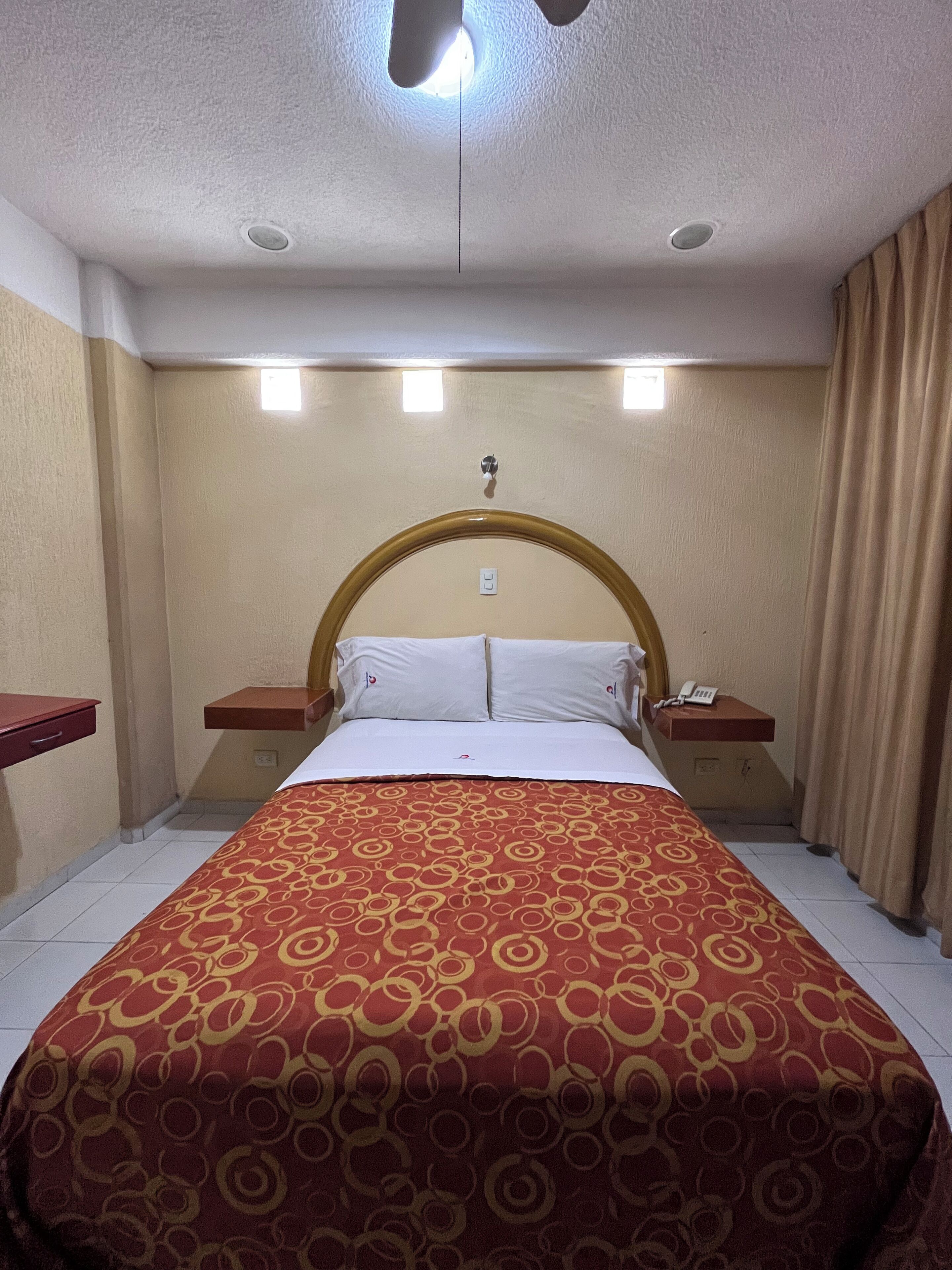 Una habitación de hotel con una cama grande, dos mesitas de noche, un cabecero y una ventana con cortinas.
