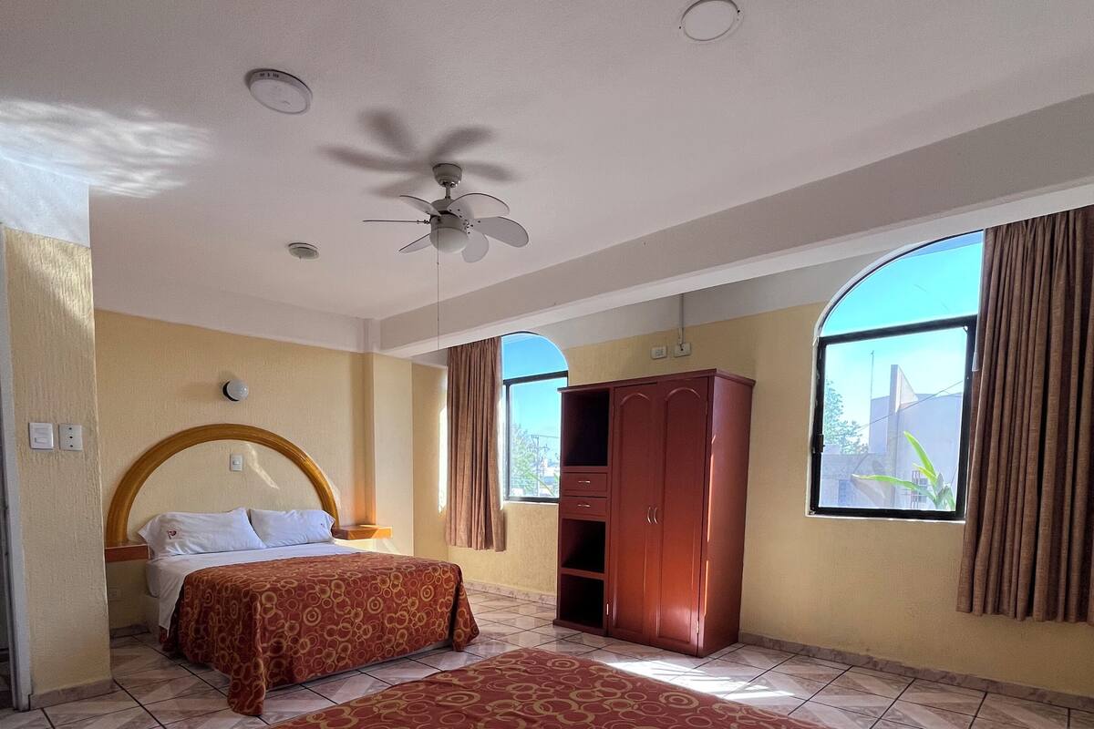 Habitación de hotel con una cama grande, una cama más pequeña, un armario, un ventilador de techo y una ventana con cortinas.