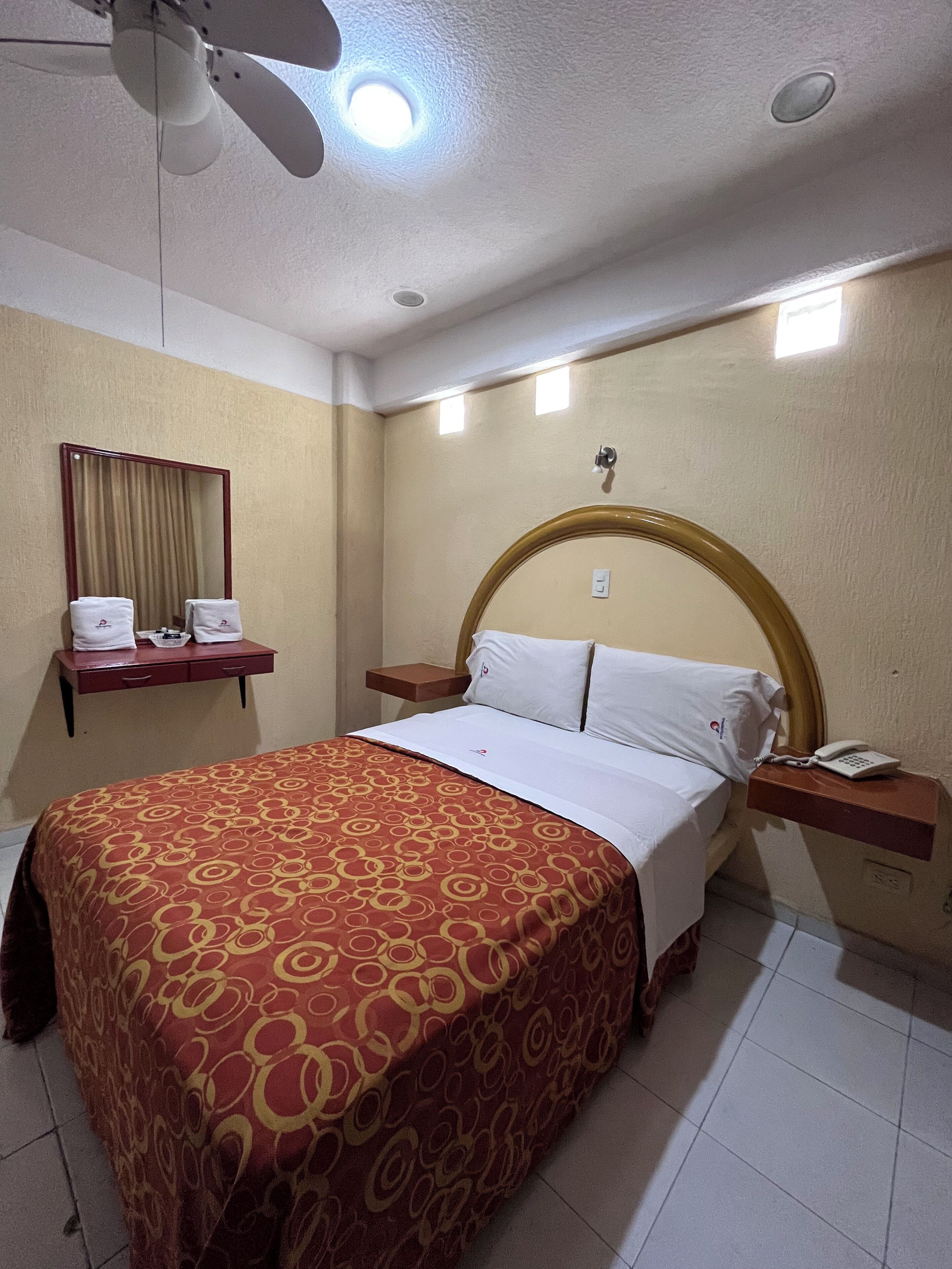 Una habitación de hotel con cama, mesita de noche con teléfono, espejo y ventilador de techo.