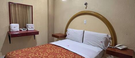 Una habitación de hotel con cama, mesita de noche con teléfono, espejo y ventilador de techo.