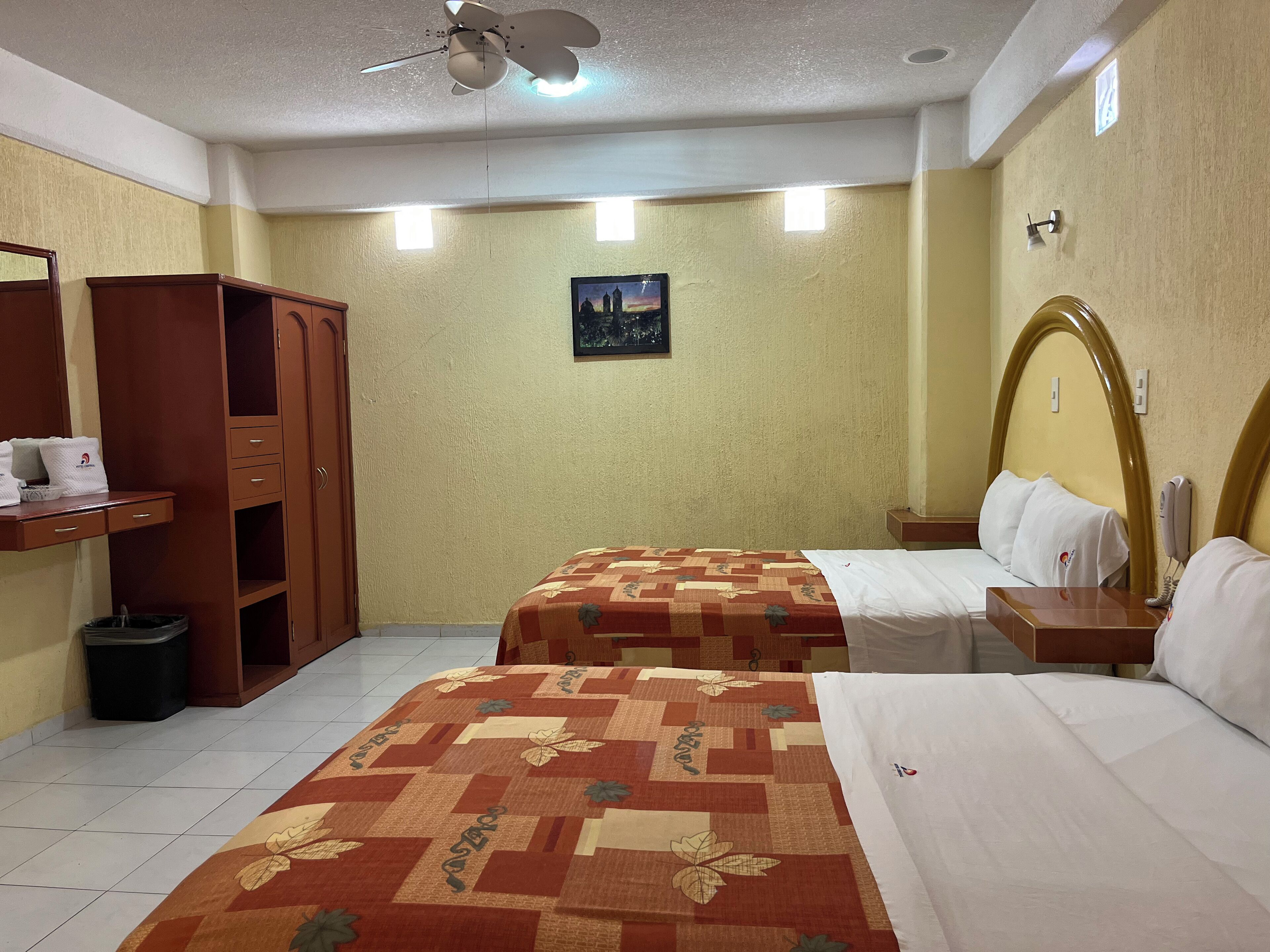 A hotel room with two beds, a wardrobe, a trash bin, a ceiling fan, and a small painting on the wall.
