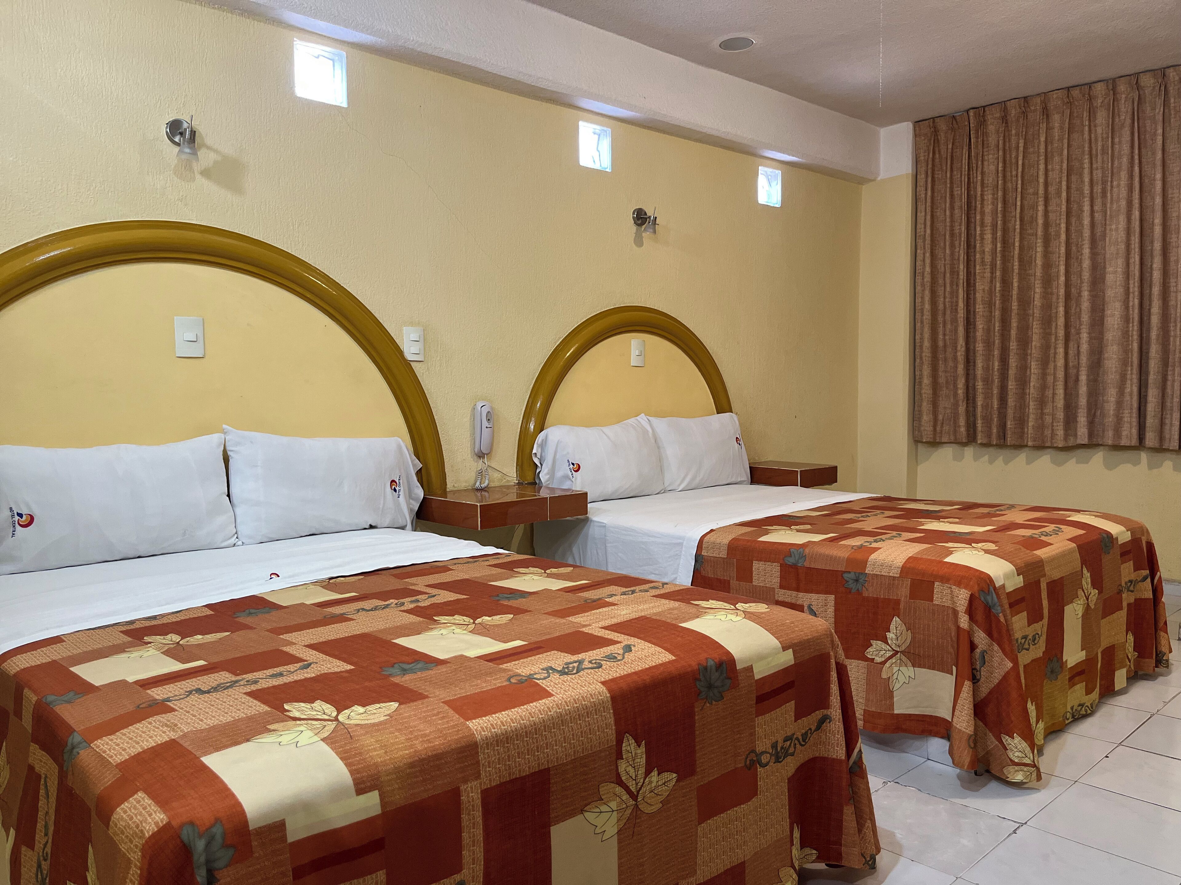 Habitación de hotel con dos camas, cada una con cabecero de madera, ropa de cama blanca y colcha con estampado de hojas. Se observa una mesita de noche con un teléfono y una lámpara pequeña. La habitación tiene una pared color beige y una ventana con cortinas.