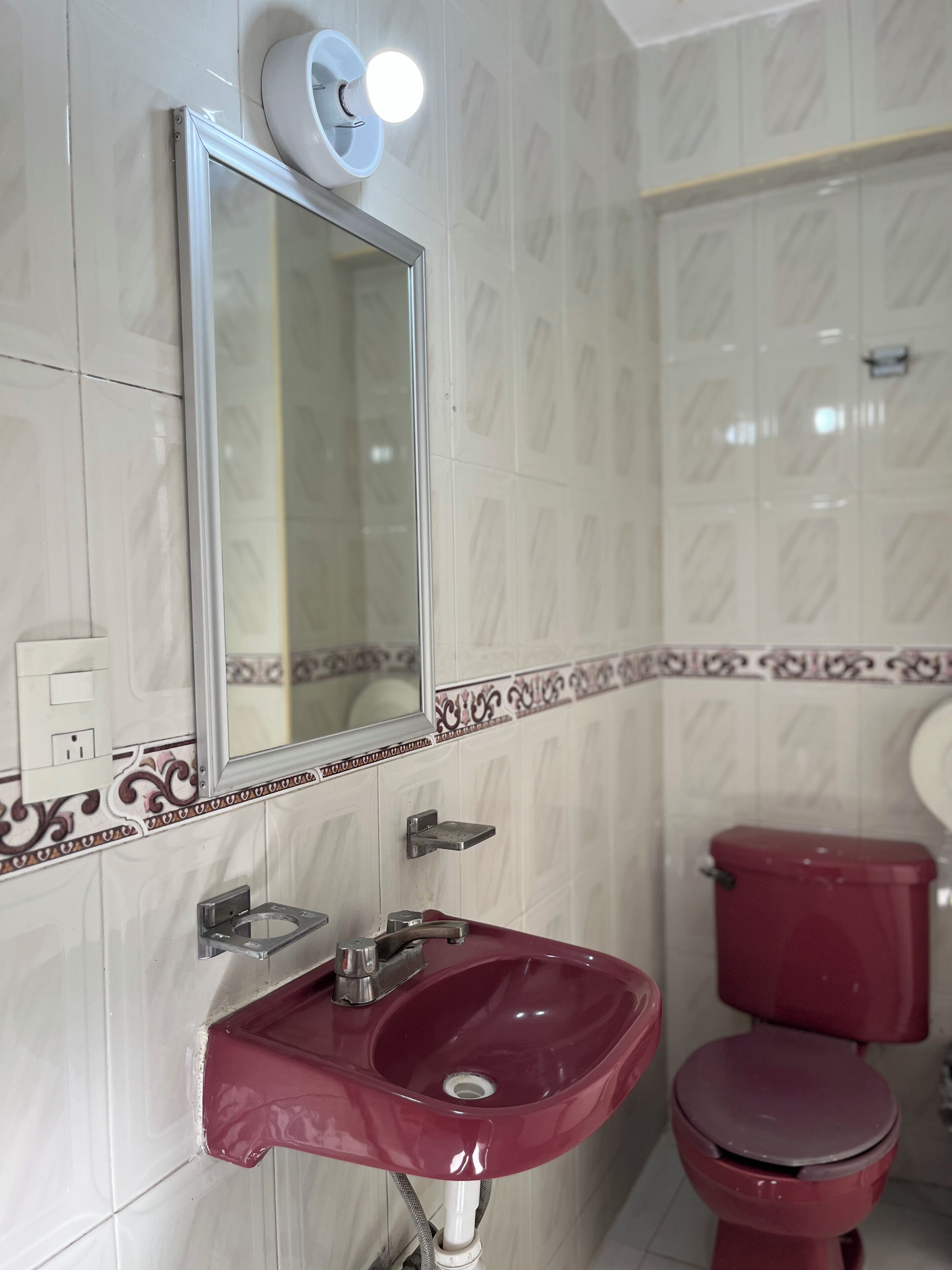 A bathroom with a white tiled wall, a red sink, a mirror, a toilet, and a wall-mounted light fixture.