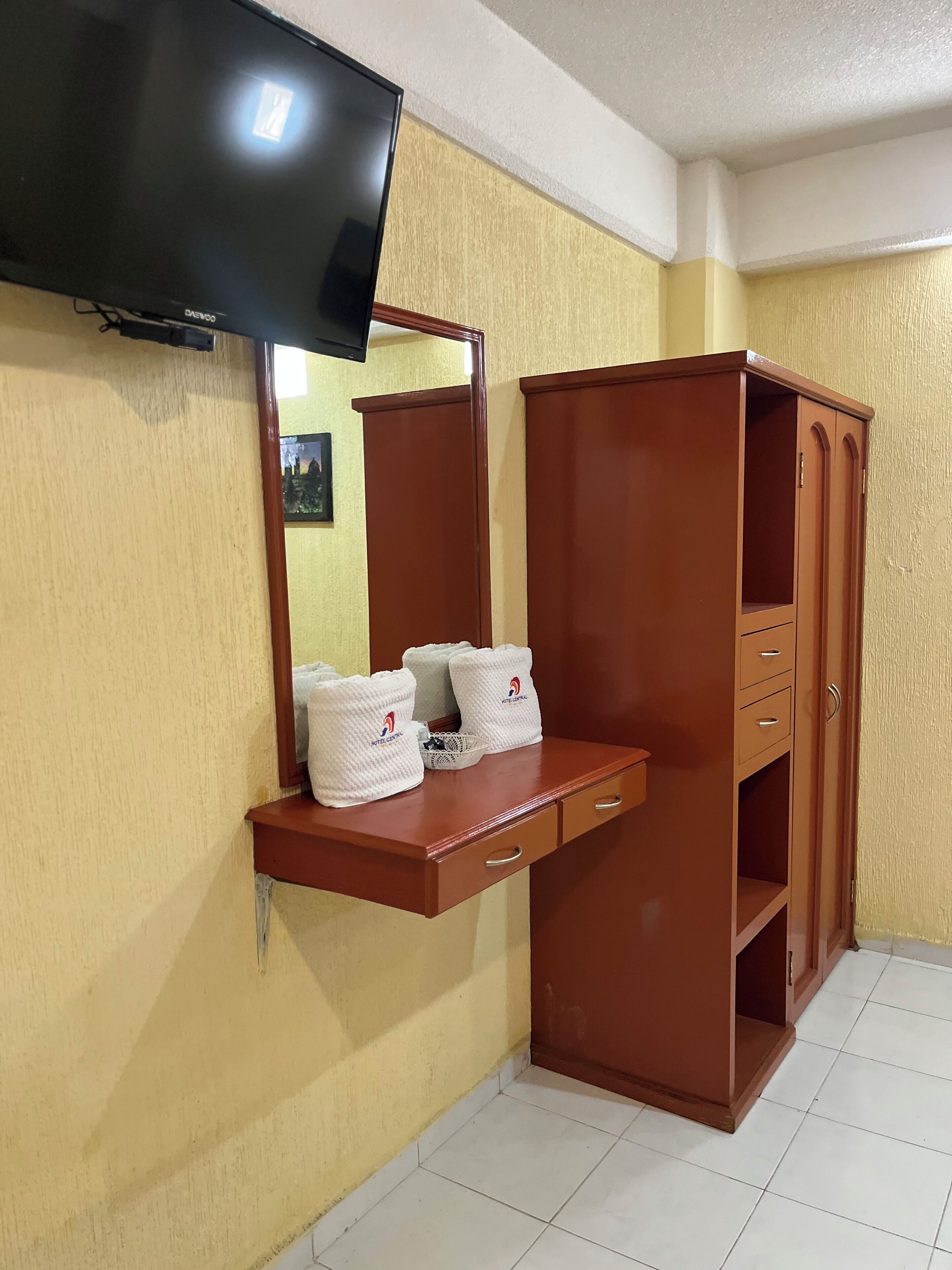 A hotel room with a wooden vanity, a flat-screen TV mounted on the wall, and a large mirror. There are towels and a basket on the vanity.