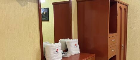 A hotel room with a wooden vanity, a flat-screen TV mounted on the wall, and a large mirror. There are towels and a basket on the vanity.