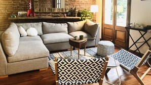A cozy living room with a grey sectional sofa, a patterned ottoman, and a wooden coffee table. A brick wall and a large window provide natural light. - Marcel de Buenos Aires (Buenos Aires)