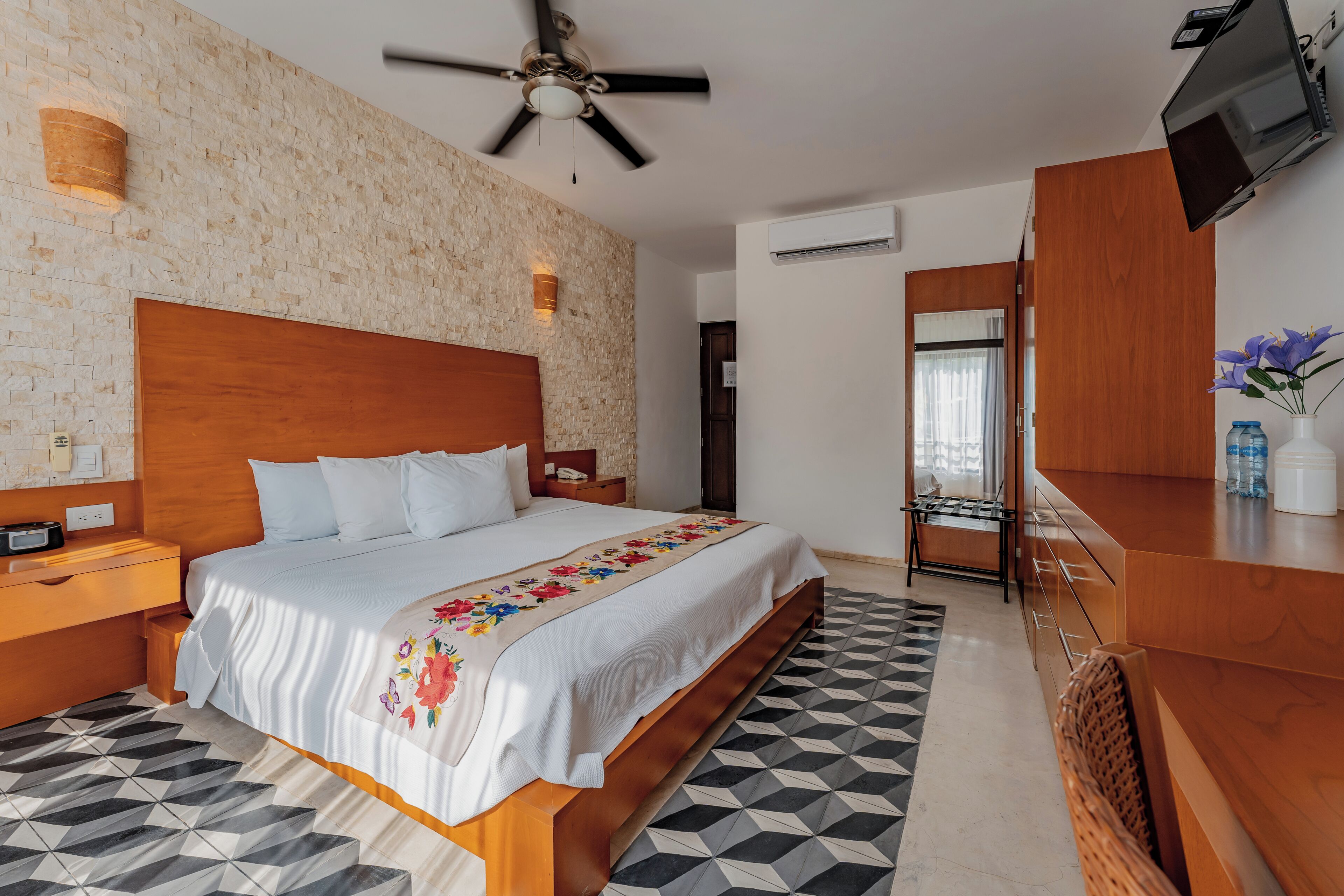 A hotel room with a large bed, wooden headboard, a ceiling fan, a TV mounted on the wall, and a geometric patterned floor.
