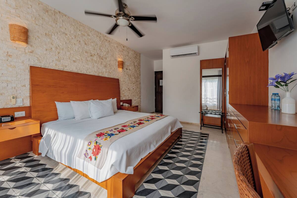 Une chambre d’hôtel dotée d’un grand lit, d’une tête de lit en bois, d’un ventilateur de plafond, d’un téléviseur fixé au mur et d’un sol orné de motifs géométriques.
