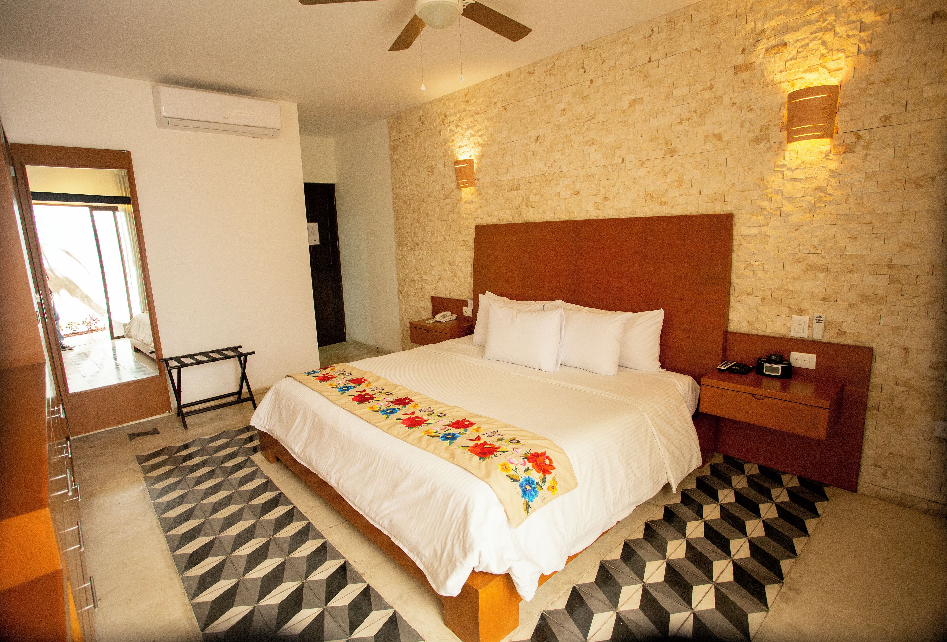Quarto de hotel com uma cama grande, cabeceira de madeira, mesa de cabeceira, luminária na parede e tapete com padrão geométrico.