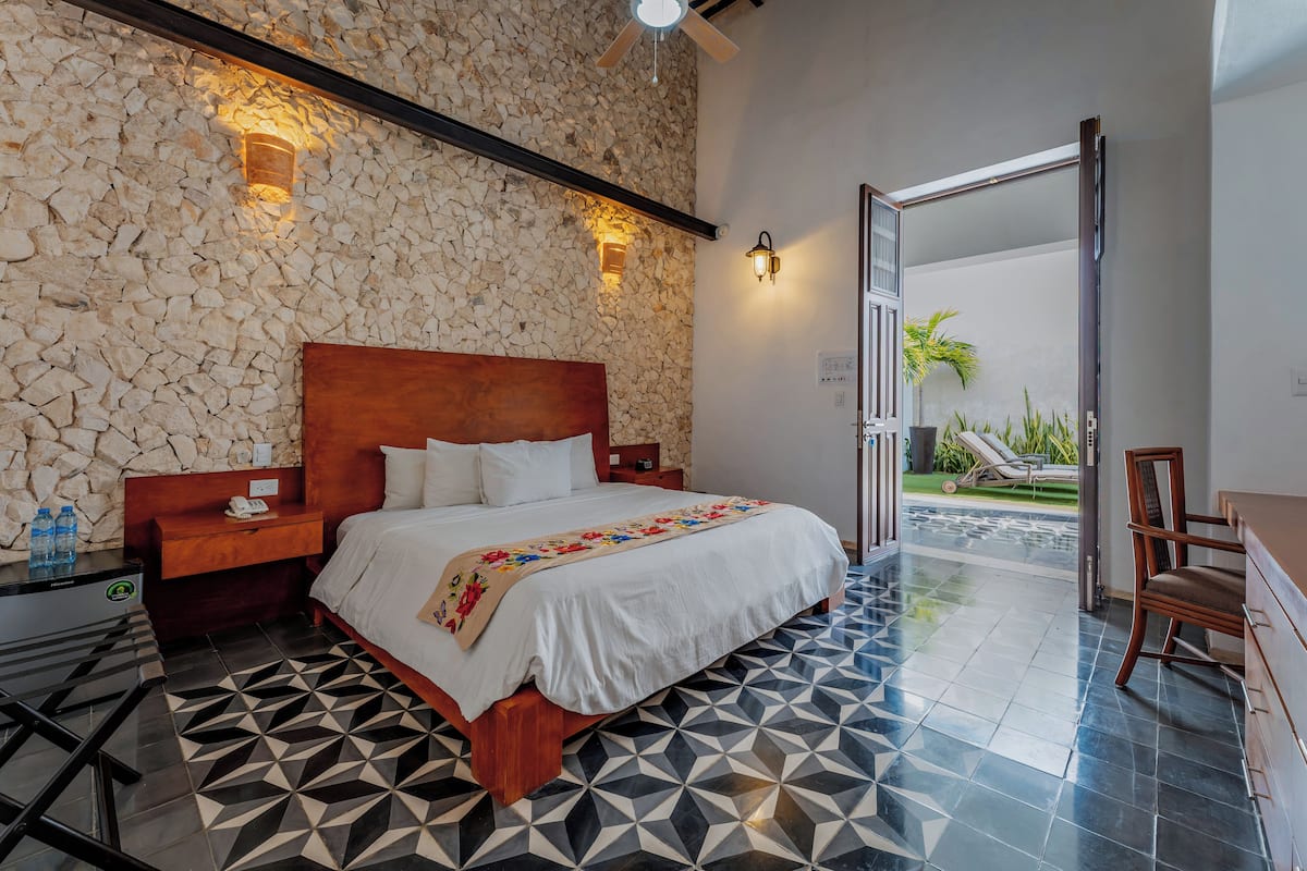 Une chambre d’hôtel avec un mur en pierre, un lit avec une tête de lit en bois, une table de chevet, une commode, une chaise et une vue sur l’extérieur.