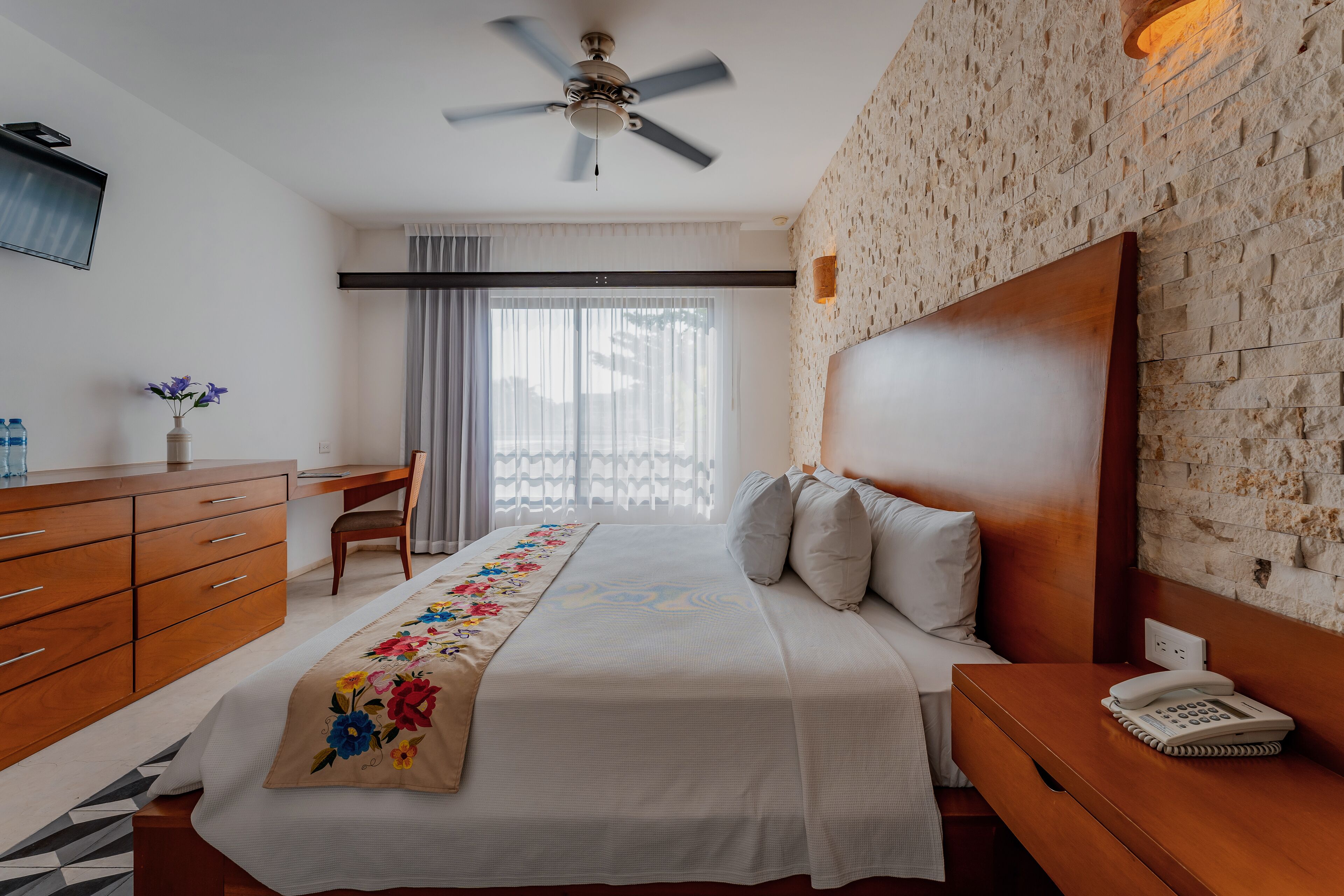 A hotel room with a bed, wooden dresser, desk, and a ceiling fan.