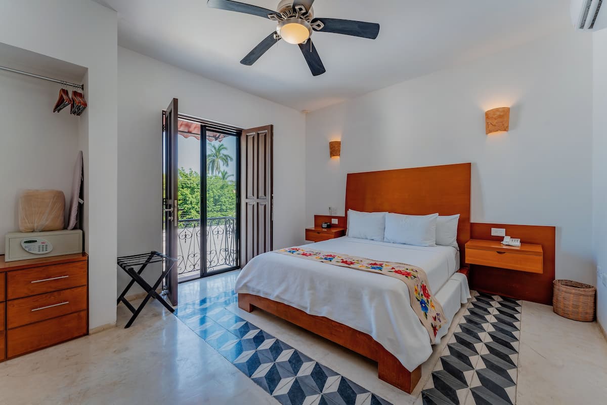 Une chambre moderne avec un grand lit, une tête de lit en bois, des tables de chevet, un ventilateur de plafond et une vue sur l’extérieur, accessible par une porte-fenêtre coulissante.