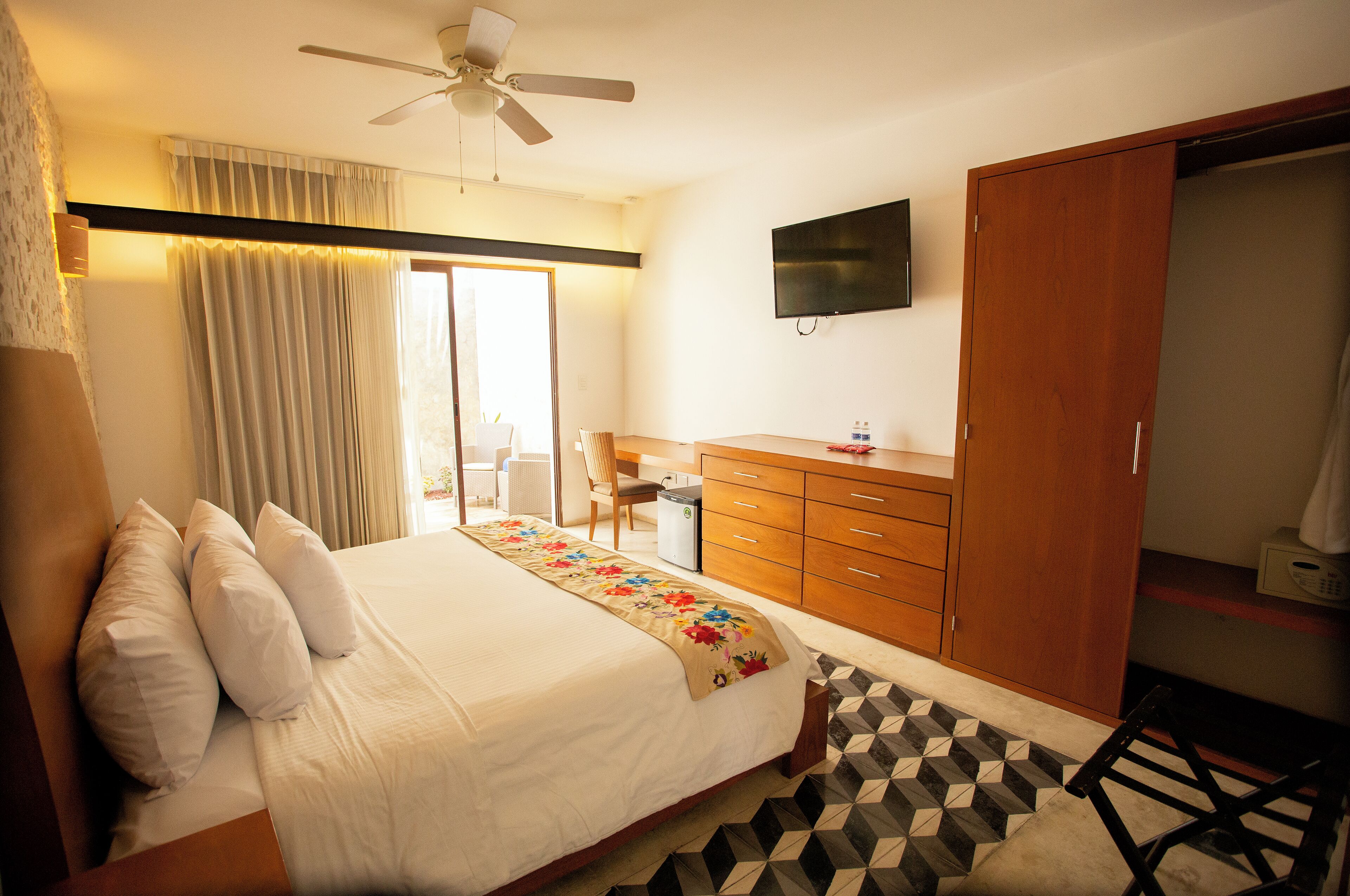 Habitación de hotel con cama, escritorio, silla, armario, televisor y ventilador de techo.