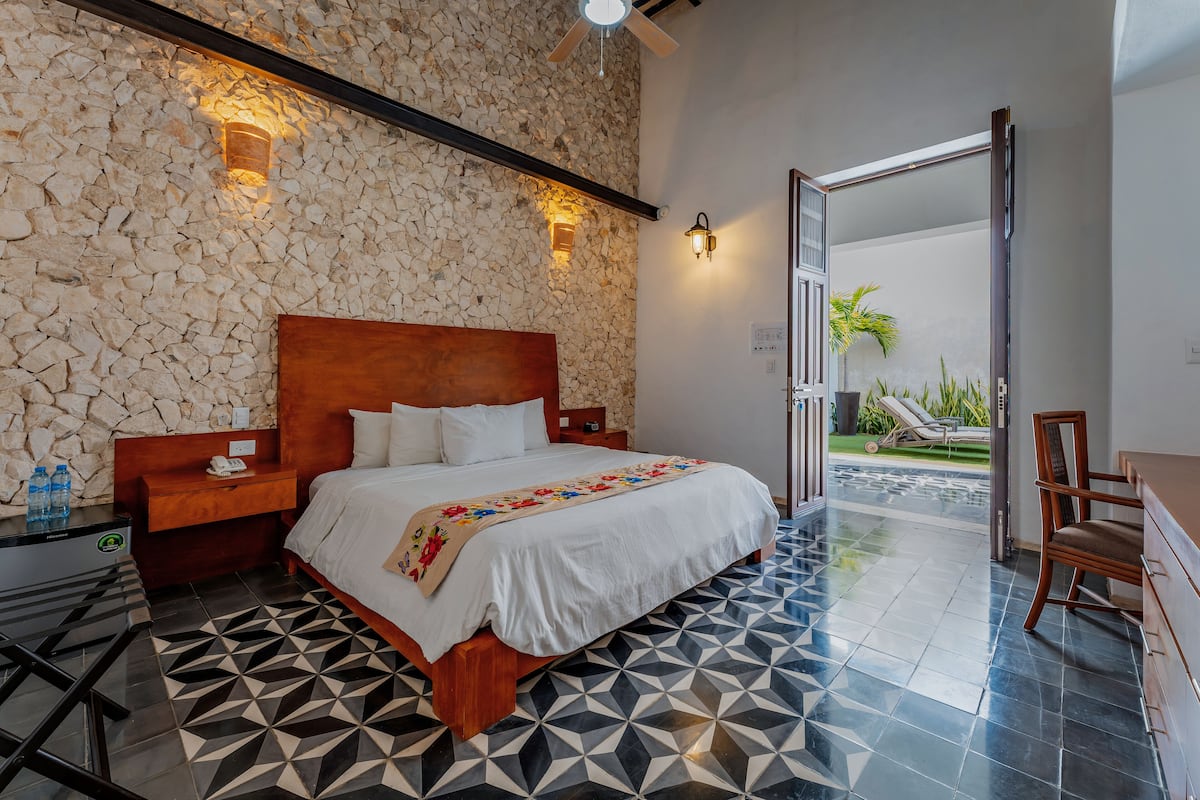 Une chambre d’hôtel avec un mur en pierre, un lit avec une tête de lit en bois, une table de chevet, une commode, une chaise et une vue sur l’extérieur.