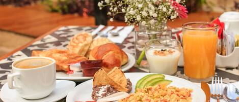 A breakfast table with croissants, a cup of coffee, a glass of orange juice, and a plate of scrambled eggs with avocado and tomatoes.