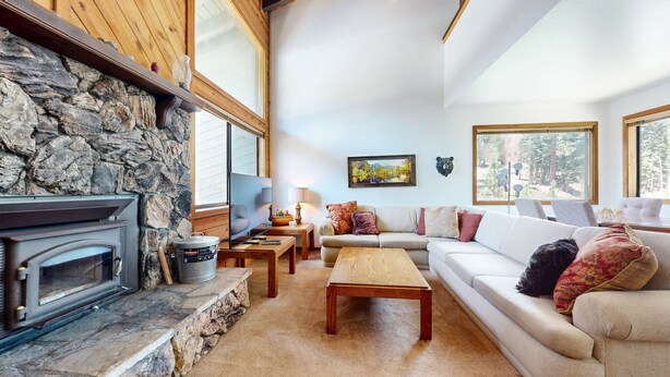 A cozy living room with a stone fireplace, a sofa set, a coffee table, and a large window offering a view of trees. - Rainbow S 4 3 Bedroom Condo by RedAwning (Mammoth Lakes)