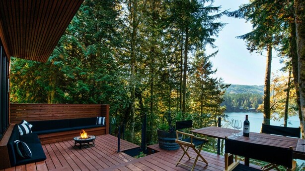 Una terraza de madera con un pozo de fuego, asientos y una mesa con una botella, con vista a un bosque y un lago.