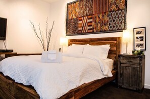 Una cama bien hecha con edredón blanco y almohadas, cabecera de madera, mesita de noche con lámpara y una obra de arte colgada en la pared.