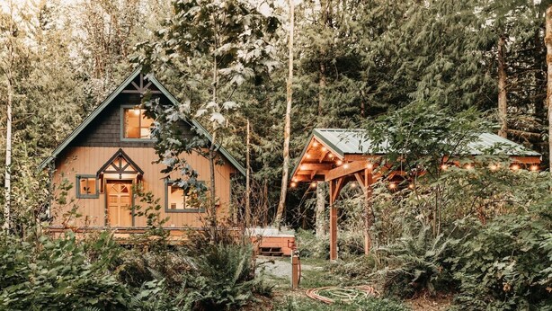 A rustic cabin with a steep gabled roof, surrounded by dense forest.