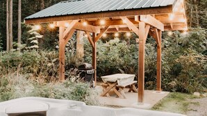 A wooden gazebo with string lights, a hot tub, and a barbecue grill in a garden setting.