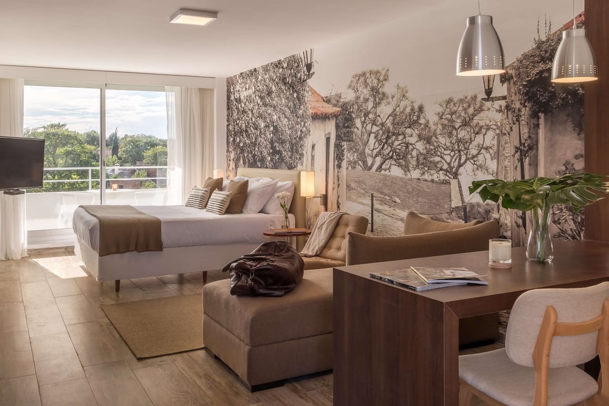 A modern hotel room with a bed, sofa, desk, and a large window offering a view of trees and buildings.