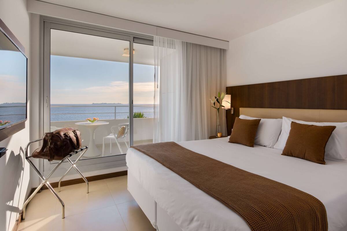 A hotel room with a large bed, a view of the sea, a balcony with a table and chairs, and a chair with a bag on it.