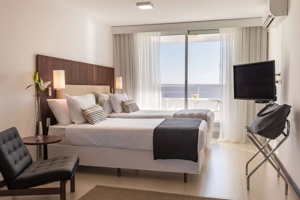 A hotel room with two beds, a TV, a chair, and a view of the sea.