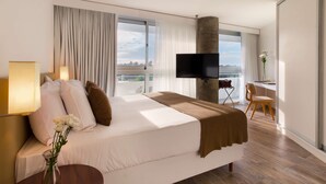 A modern hotel room with a large bed, a flat-screen TV, a desk with a chair, and a view of the city through large windows. - Dazzler by Wyndham Colonia (Colonia del Sacramento)