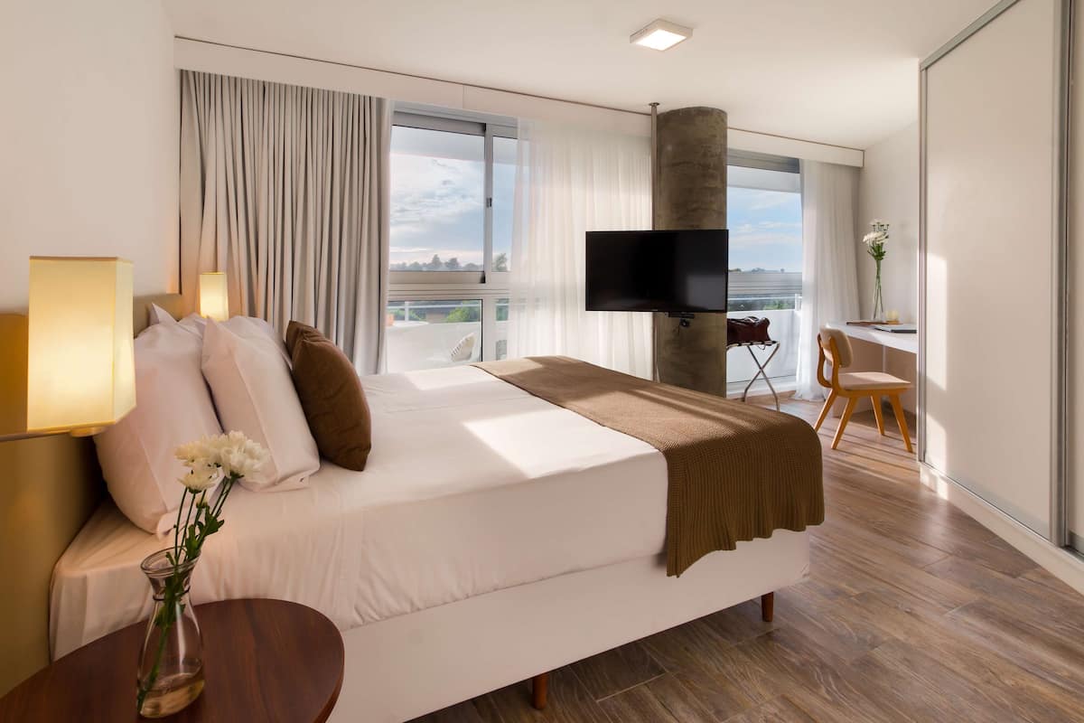A modern hotel room with a large bed, a flat-screen TV, a desk with a chair, and a view of the city through large windows.