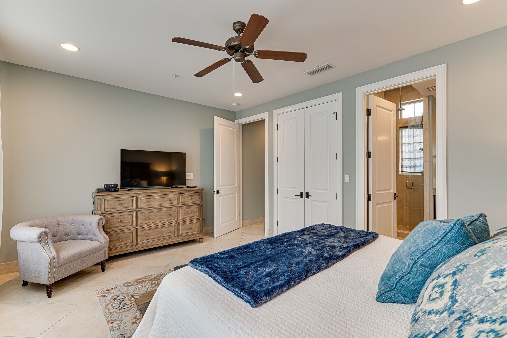 Une chambre à coucher comprenant un lit, un ventilateur de plafond, une commode, un téléviseur et une porte donnant accès à une salle de bain.