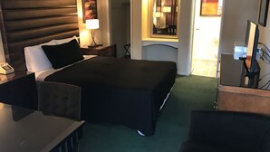 Habitación de hotel con una cama grande, un escritorio, una silla, una mesita de noche con lámpara, un espejo y vistas al exterior.