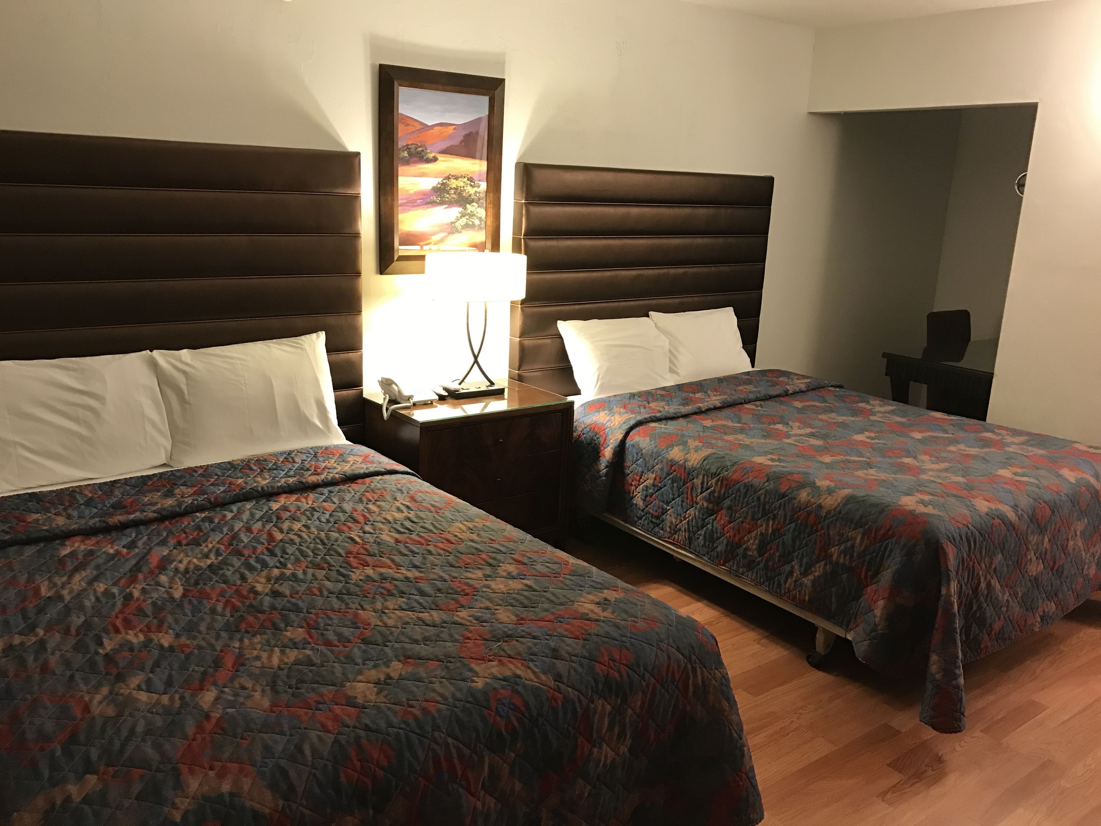 Quarto de hotel com duas camas, uma mesa de cabeceira com abajur, um quadro na parede e uma escrivaninha com cadeira.