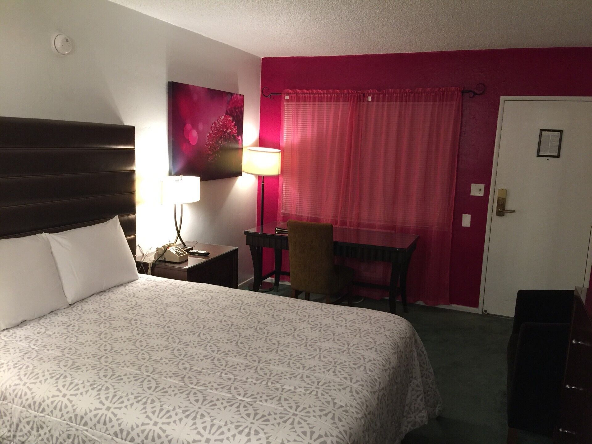 A hotel room with a bed, a desk, a chair, a lamp, and a door.