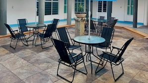An outdoor patio area with round tables and chairs, blue umbrellas, and a white building with columns in the background. - Casa Porfirio (Oaxaca)