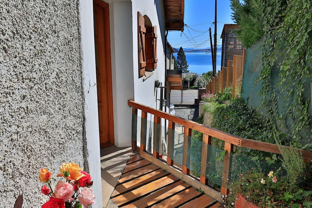 Un balcón de madera con barandilla metálica, una pared blanca y vista al mar.