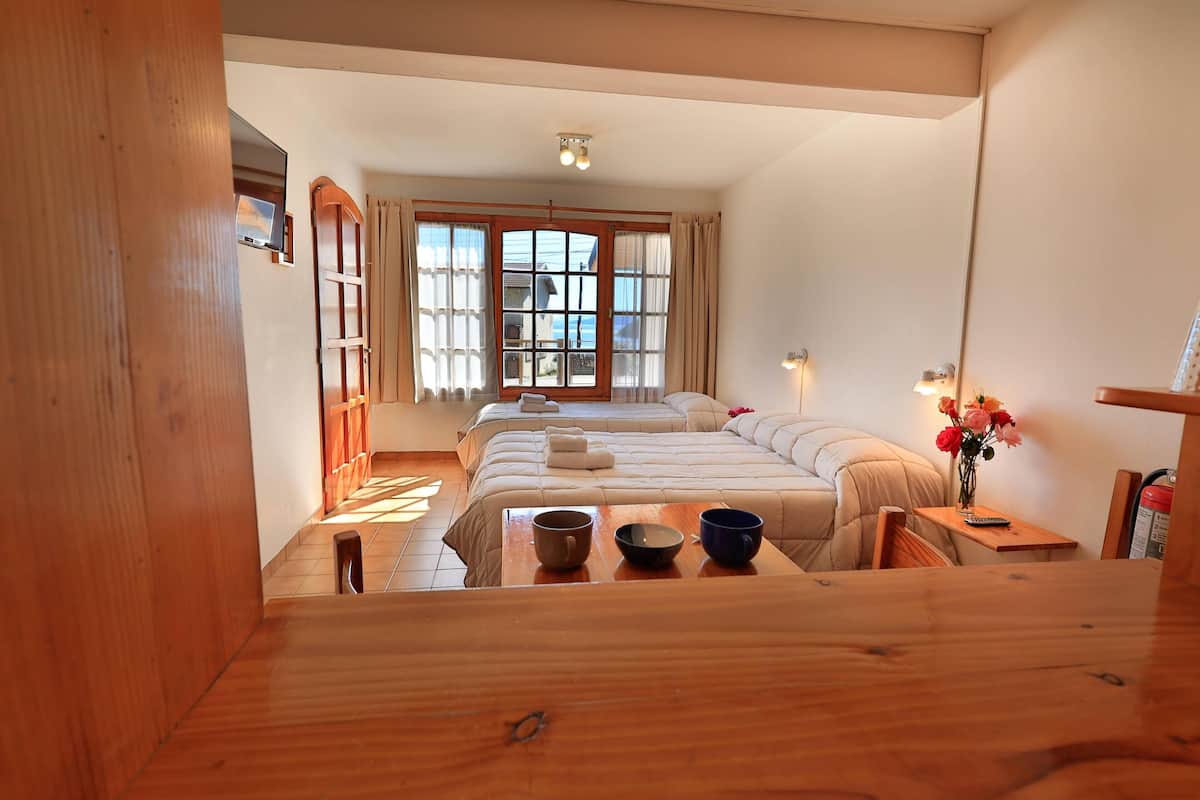 Habitación de hotel con una cama grande, una mesa de madera con tazas, una mesita con un florero y una ventana con cortinas.