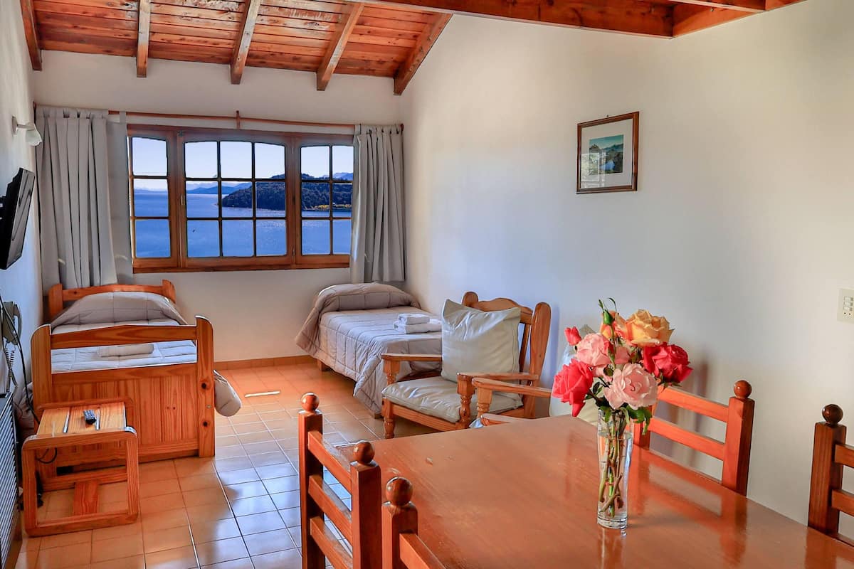 Una habitación con dos camas de madera, una mesa de madera con sillas, una ventana con cortinas y un jarrón con flores.