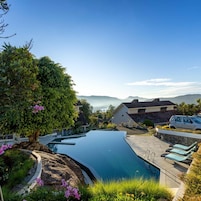 Una piscina con vistas a una cadena montañosa, rodeada de vegetación y un edificio blanco.