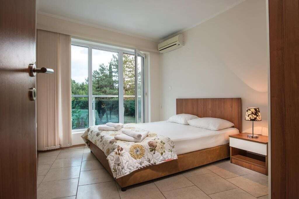 A bedroom with a large bed, a nightstand, a window with curtains, and an air conditioning unit.